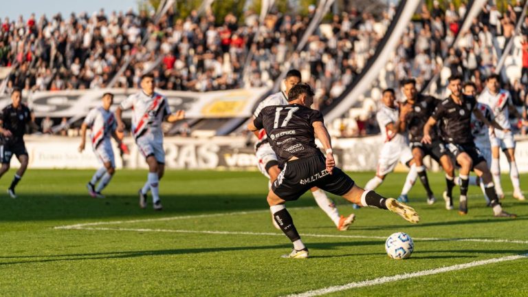 Jogadores disputam uma partida de futebol diante de um estádio lotado em um dia ensolarado.