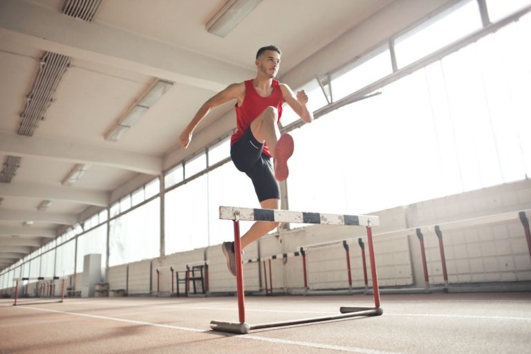 Atleta masculino salta sobre uma barreira durante treino em uma pista coberta.